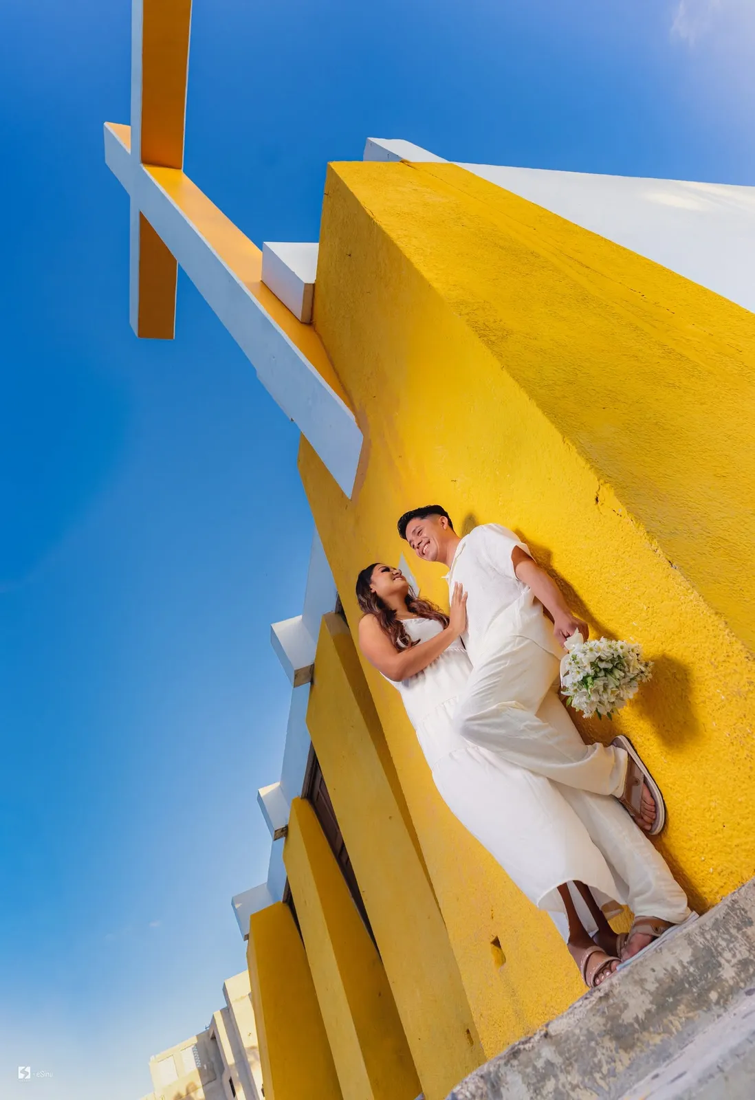 Novia con vestido blanco junto a un muro amarillo, vista desde abajo