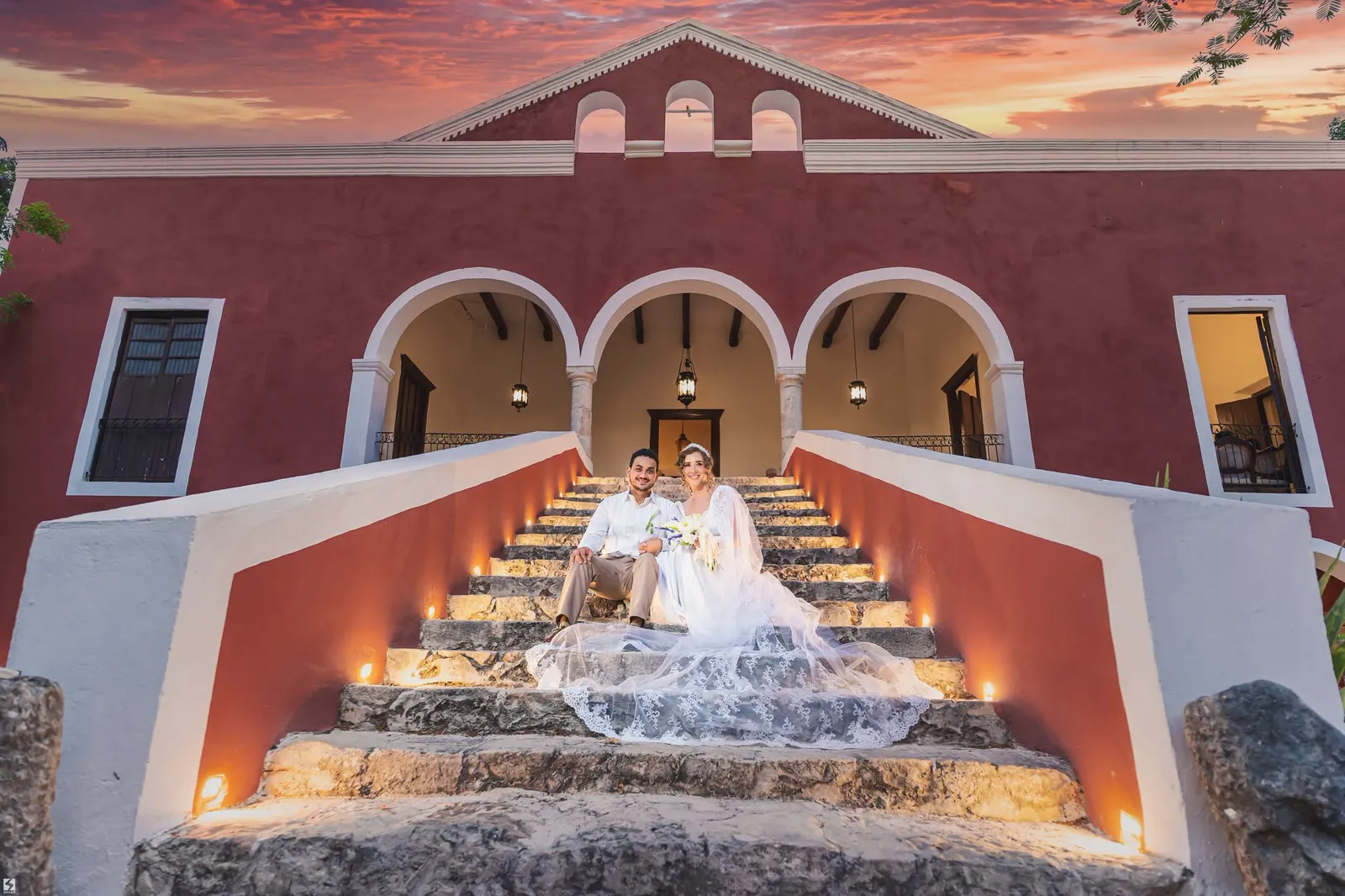 Novia sentada en la escalinata de una hacienda roja al atardecer
