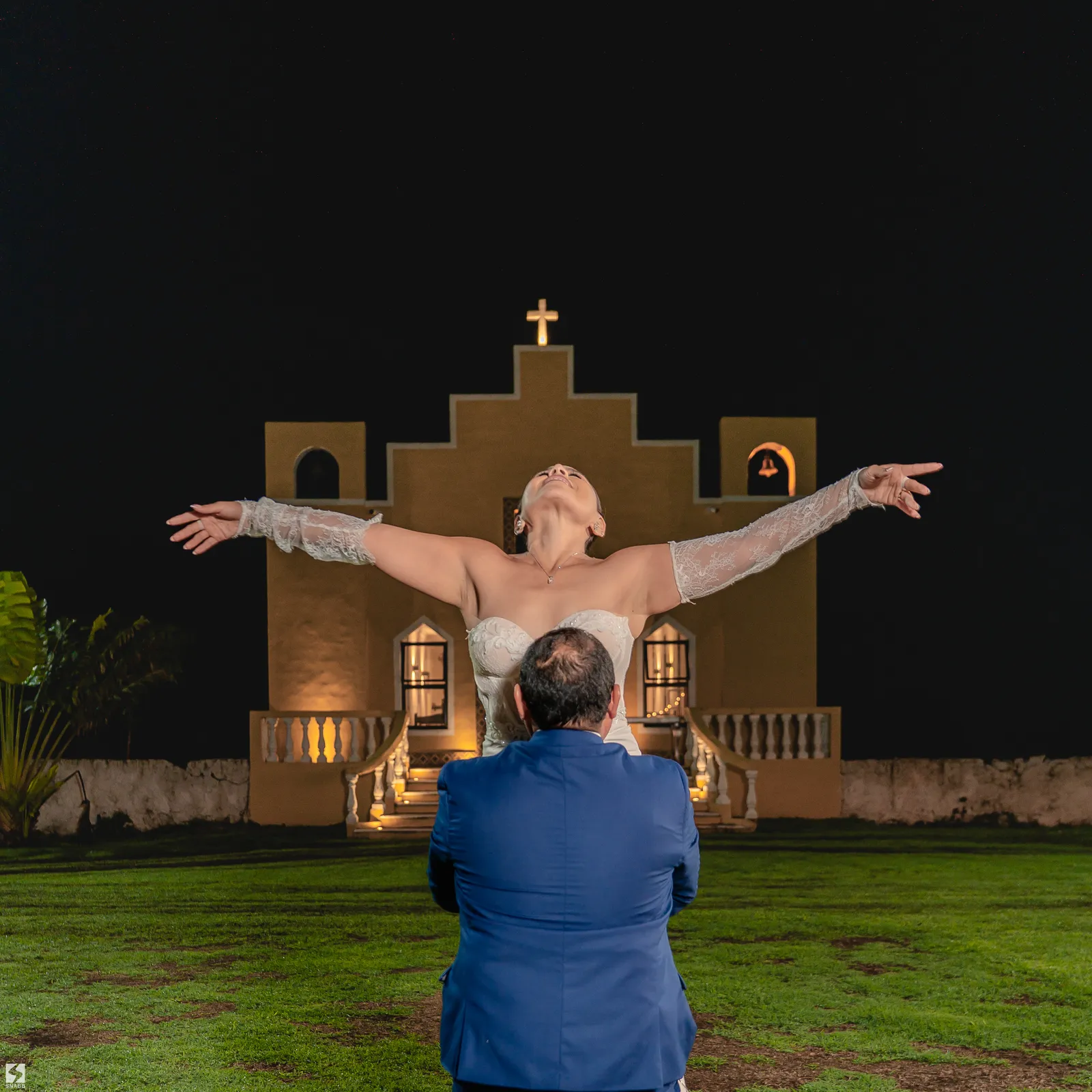 Novia con los brazos abiertos frente a una capilla iluminada de noche